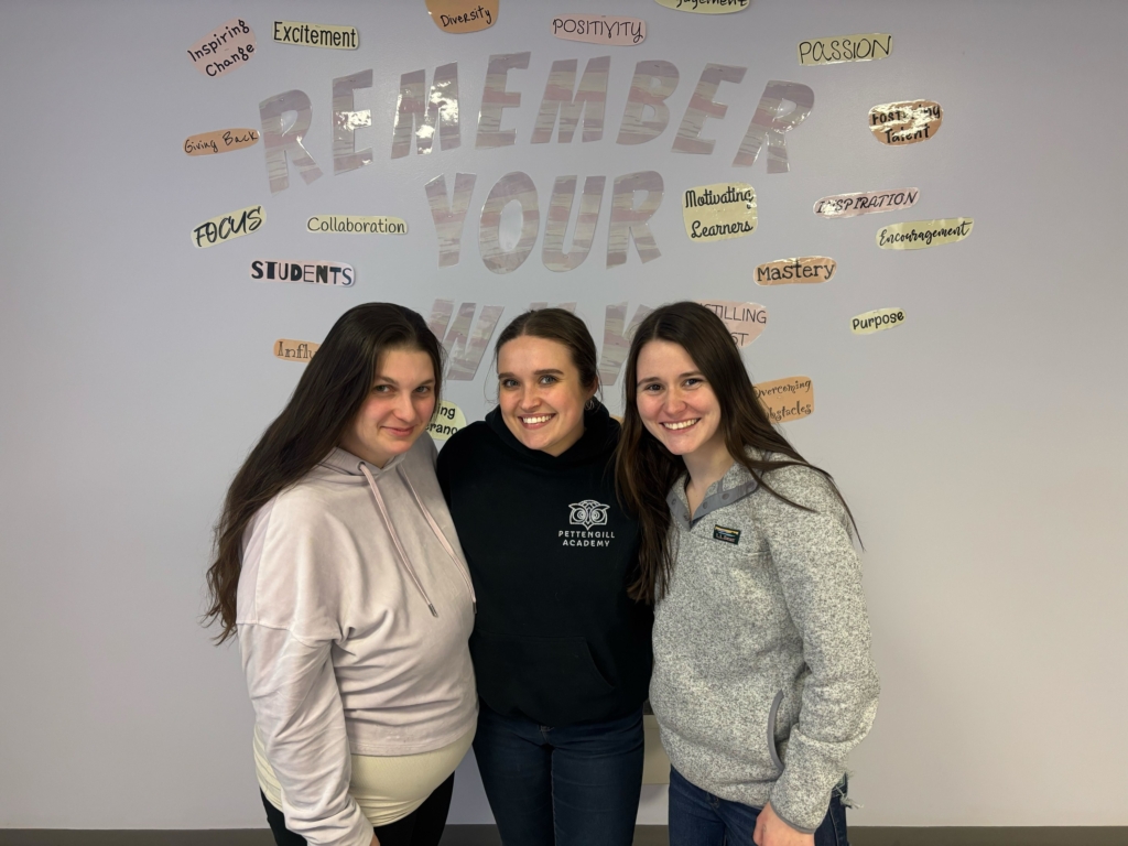 Three women embrace each other in front of an inspiring wall, highlighting themes of collaboration and positivity in their workspace.