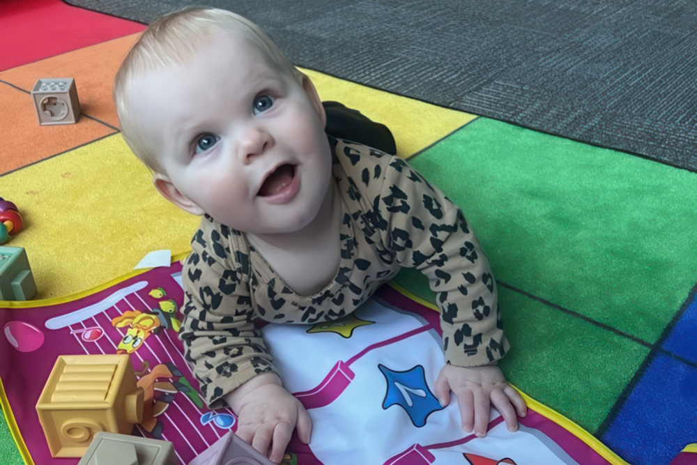 A delighted baby crawls on a colorful play mat, looking up with a big smile while surrounded by toys.