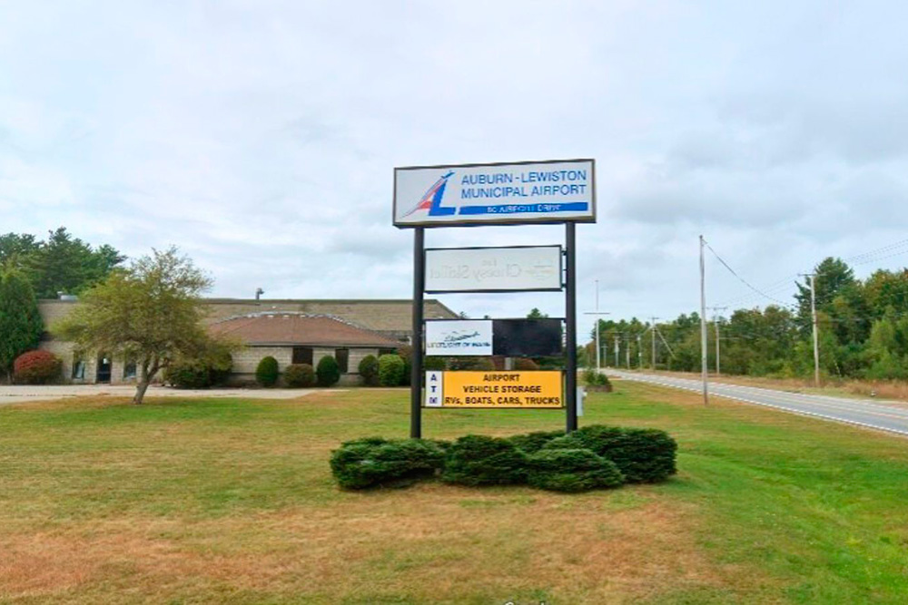 A sign for Auburn-Lewiston Municipal Airport, indicating airport services and vehicle storage, set against a cloudy sky.