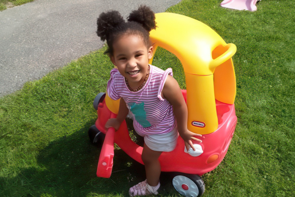 A young girl with puffy hair smiles while playing on a red and yellow toy car, enjoying a sunny day on the grass.