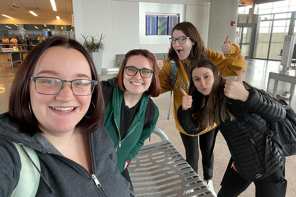 Four friends take a cheerful selfie at the office all smiling and giving thumbs up, excited for their upcoming adventure.