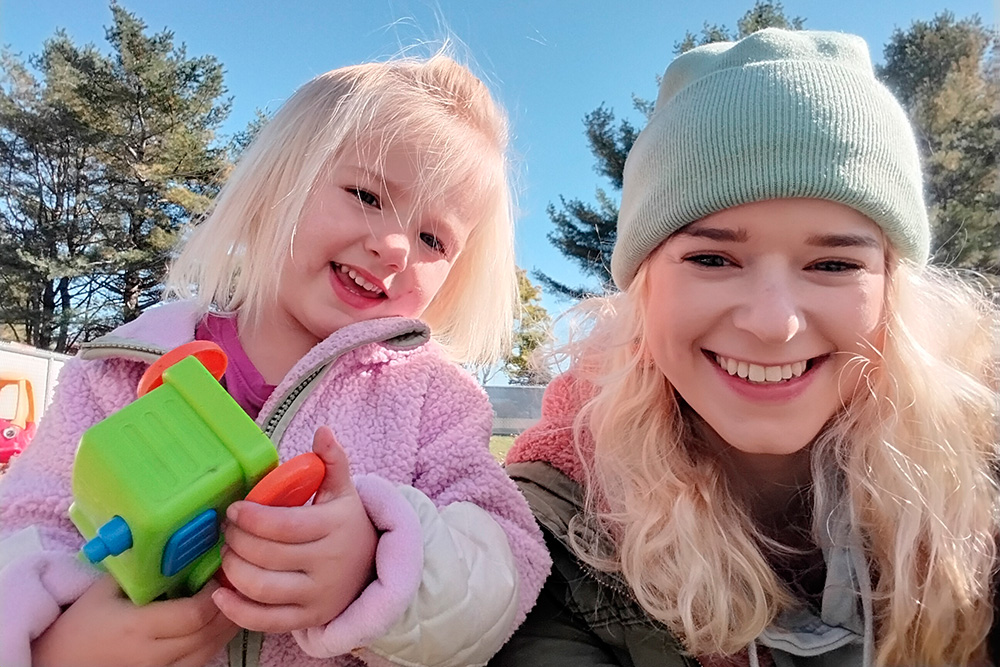 A girl holds a toy and smiles at the camera with her parent beside her, both enjoying a sunny day outdoors.