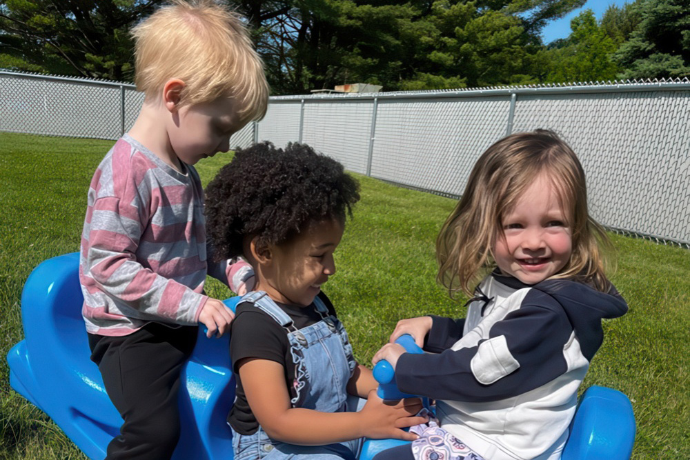 Three kids play on a blue ride-on toy outdoors, with one smiling at the camera while the others are engaged in fun.