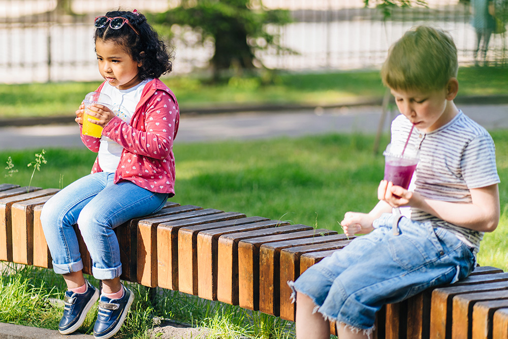 Two children sit on a bench, each enjoying drinks, absorbed in their own world on a sunny day.